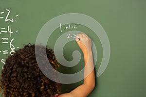 Schoolgirl solving maths formula on green board in classroom