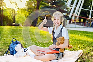 Schoolgirl student happy smiling showing thumb up sits on a blanket in the park on a sunny day. the teenager is holding a bear toy