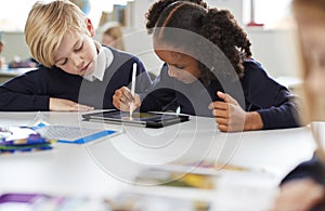 Schoolgirl using a tablet and stylus sitting with a boy at a desk in a primary school classroom, selective focus