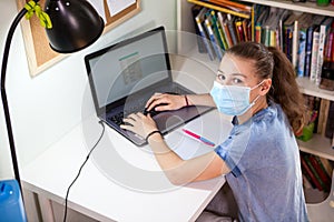 Schoolgirl working on laptop while sitting at home, child wearing medical mask suring self-isolation