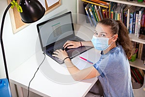 Schoolgirl working on laptop while sitting at home, child wearing medical mask suring self-isolation photo