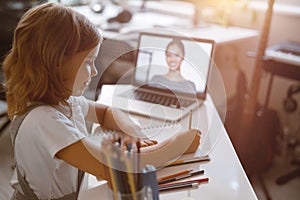 Schoolgirl writes listening to teacher at videocall via modern laptop at table in light room