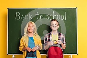 Schoolkids with backpacks standing near chalkboard with back to school lettering