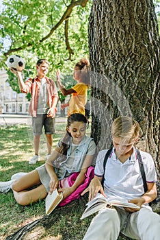 Schoolkids sitting under tree and reading books near multicultural friends