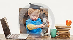 Science, Biology, Experiment, Education. Thoughtful smart boy in graduation hat working with microscope and laptop.