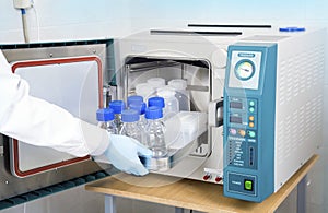 Scientific researcher hand in blue glove inserts tray with glass and plastic bottles into the autoclave