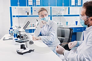scientific researchers in medical masks and goggles having discussion at workplace with microscope
