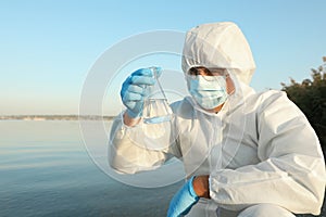 Scientist in chemical protective suit with conical flask taking sample from river for analysis