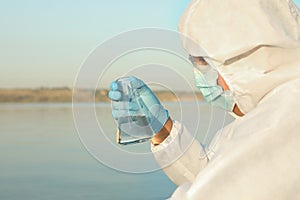 Scientist in chemical protective suit with conical flask taking sample from river for analysis