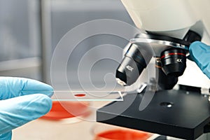 Scientist examining sample of red liquid on slide under microscope in laboratory, closeup