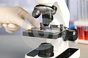 Scientist examining sample of red liquid on slide under microscope in laboratory, closeup