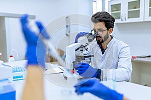 Scientist in glasses reserching microorganisms in microscope, working with unrecognizable colleague