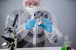 Scientist in hazmat suit and goggles holding vaccine bottle and magnifying glass, looking and research. Epidemiology virus test.