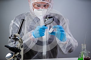 Scientist in hazmat suit and goggles holding vaccine bottle and magnifying glass, looking and research. Epidemiology virus test.