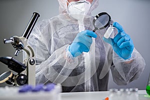 Scientist in hazmat suit and goggles holding vaccine bottle and magnifying glass, looking and research. Epidemiology virus test.