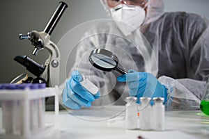 Scientist in hazmat suit and goggles holding vaccine bottle and magnifying glass, looking and research. Epidemiology virus test.