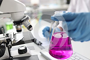 Scientist holding flask with pink liquid in front of microscope closeup