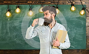 Scientist holds book and drinks coffee, chalkboard on background, copy space. Man with beard on calm face in classroom