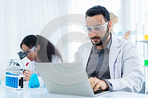 Scientist in lab coat and safety glasses holding flask with blue solution at modern laboratory. Take note on the laptop for