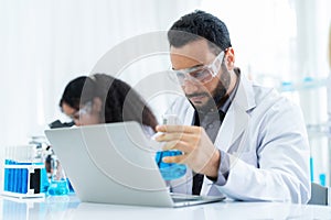 Scientist in lab coat and safety glasses holding flask with blue solution at modern laboratory. Take note on the laptop for