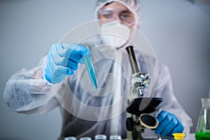 Scientist or medic in protective overalls, takes test tube with blue reagent, laboratory glassware containing chemical