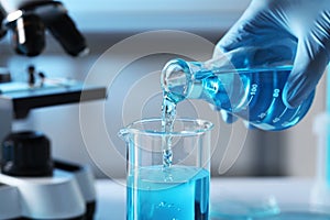 Scientist pouring light blue liquid from flask into beaker in laboratory, closeup