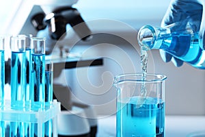 Scientist pouring light blue liquid from flask into beaker near test tubes in laboratory, closeup