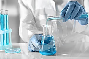 Scientist pouring light blue liquid from test tube into flask in laboratory, closeup