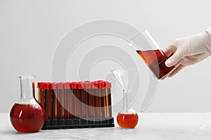 Scientist pouring liquid from beaker into conical flask on white table against light background, closeup