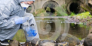 A scientist in a protective suit and mask, collects a sample of water in test tubes, against the background of drain pipes, there