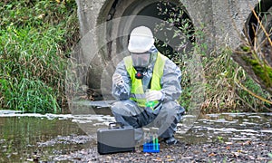 A scientist, in a protective suit and mask, takes the liquid from the river in test tubes, for a sample, against the background of