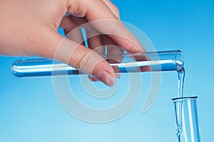 Scientist`s hand holds laboratory glass test tube with pure liquid, pours into other, makes experiment in research lab, blue backg