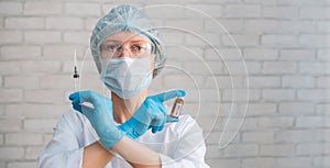 Scientist woman wearing protective suit and mask holds in crossed hands syringe and ampule. Female doctor in hospital ready for