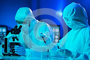 Scientists in protection suits and masks working in research lab using laboratory equipment: microscopes, test tubes