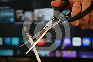 Scissors cutting through a coaxial cable with TV out of focus in background - cut cable tv concept