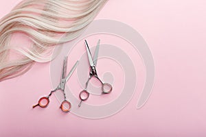 Scissors and other hairdresser`s accessories and strand of blonde hair on pink background. Flat lay with space for text.