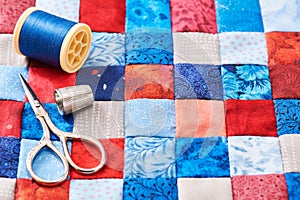 Scissors, thread and thimble lying on blue and red square pieces of fabric sewn together