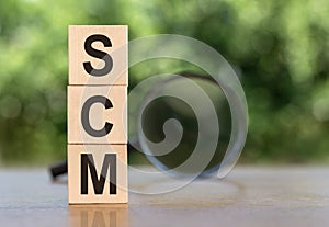 SCM Supply Chain Management abbreviation. Wooden cubes with letters, on a table with a magnifying glass. Green background.