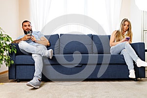 Scolded couple sitting on opposite sides of the couch using their smartphones, don't care for each other