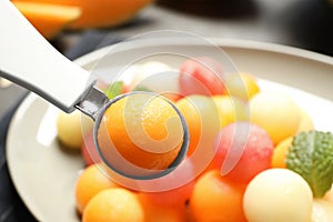 Scoop with melon ball near plate of fruit salad