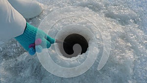 Scooping ice out of the hole during winter fishing. The hole freezes in the cold. A woman in blue gloves and white boots