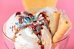 Scoops of ice cream in glass bowl with chocolate sauce, strewed sprinkles and cookies on pink background, close up