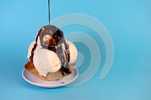 Scoops of vanilla ice cream in waffle cone bowl pouring chocolate glaze on blue background
