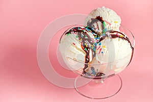 Scoops of white ice cream in glass bowl decorated chocolate topping and strewed sprinkles on pink background