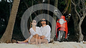 Scooter road trip. Lovely couple on red motorbike in white clothes on sand beach. People walking near the tropical palm