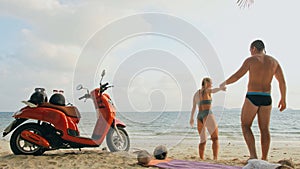 Scooter road trip. Lovely couple on red motorbike in white clothes on sand beach. People walking near the tropical palm