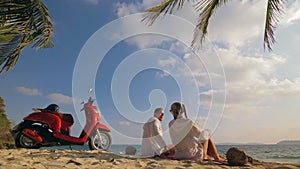 Scooter road trip. Lovely couple on red motorbike in white clothes on sand beach. People walking near the tropical palm