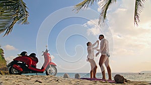 Scooter road trip. Lovely couple on red motorbike in white clothes on sand beach. People walking near the tropical palm