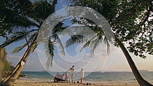 Scooter road trip. Lovely couple on red motorbike in white clothes on sand beach. People walking near the tropical palm