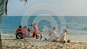 Scooter road trip. Lovely couple on red motorbike in white clothes on sand beach. People walking near the tropical palm